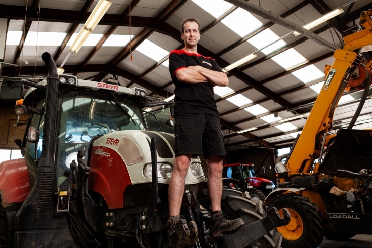Man staat op rode tractor in werkplaats.