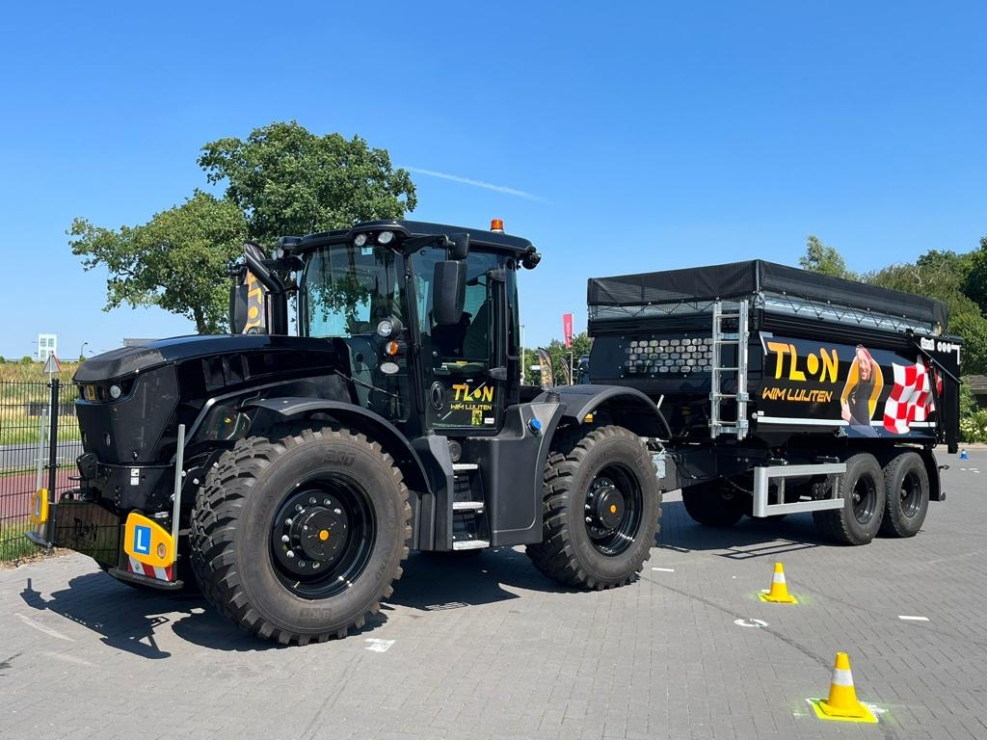 Zwarte tractor met trailer op parkeerterrein met kegels.